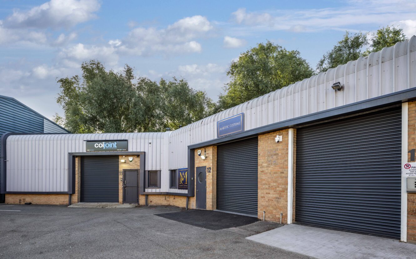 Industrial units with brick and metal exteriors, three large roller shutter doors, and signs on the buildings, set against a backdrop of trees and a partly cloudy sky.