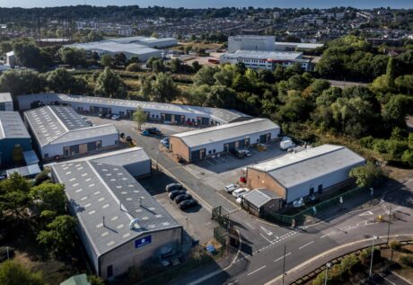 Aerial view of an industrial estate with several warehouses, parked vehicles, surrounding trees, and a residential area in the background.