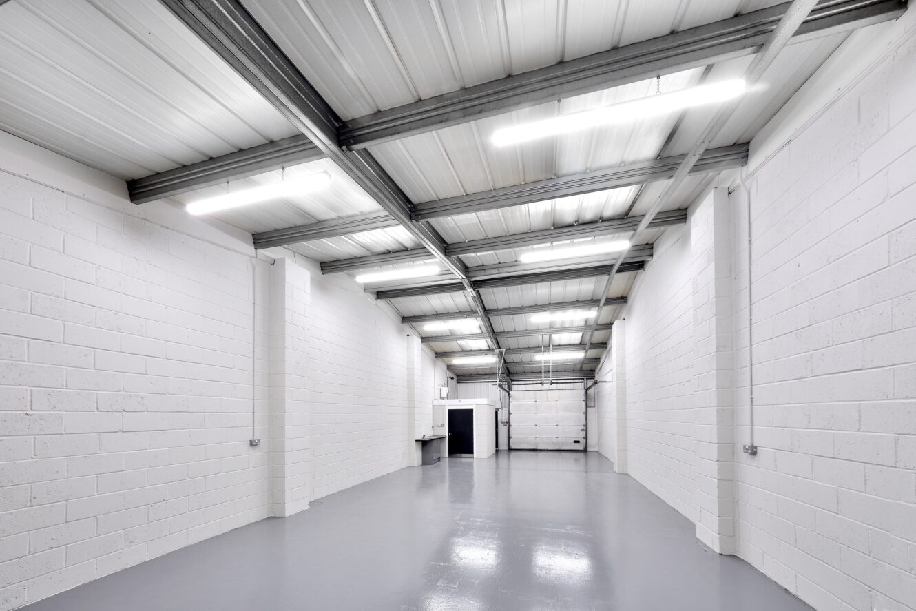 Empty industrial warehouse with white brick walls, polished gray floor, metal ceiling beams, fluorescent lighting, and a small door and sink at the far end.