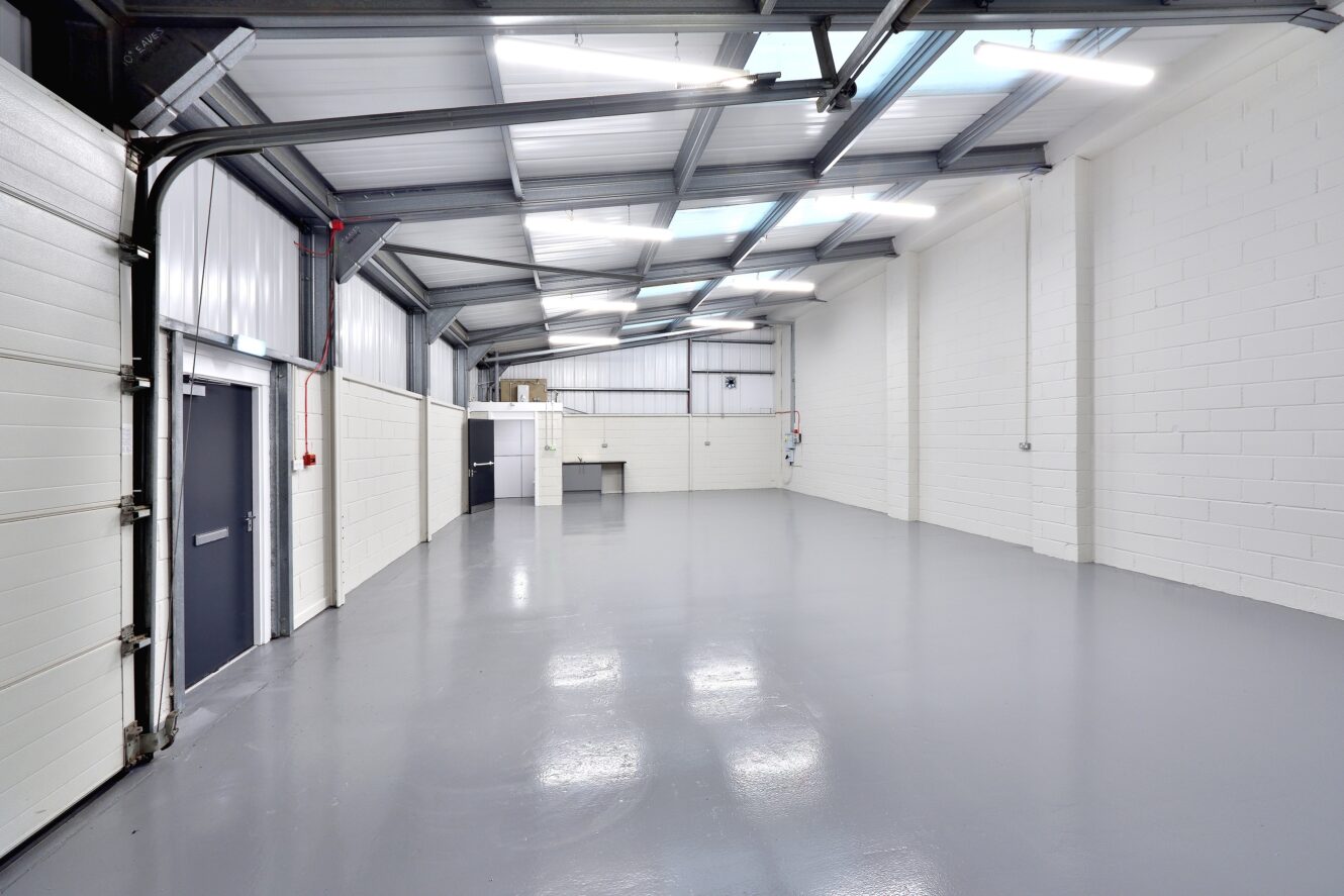 Empty industrial warehouse with white brick walls, grey epoxy floor, high ceiling with skylights, and metal beams; a closed double door and a utility sink are visible.