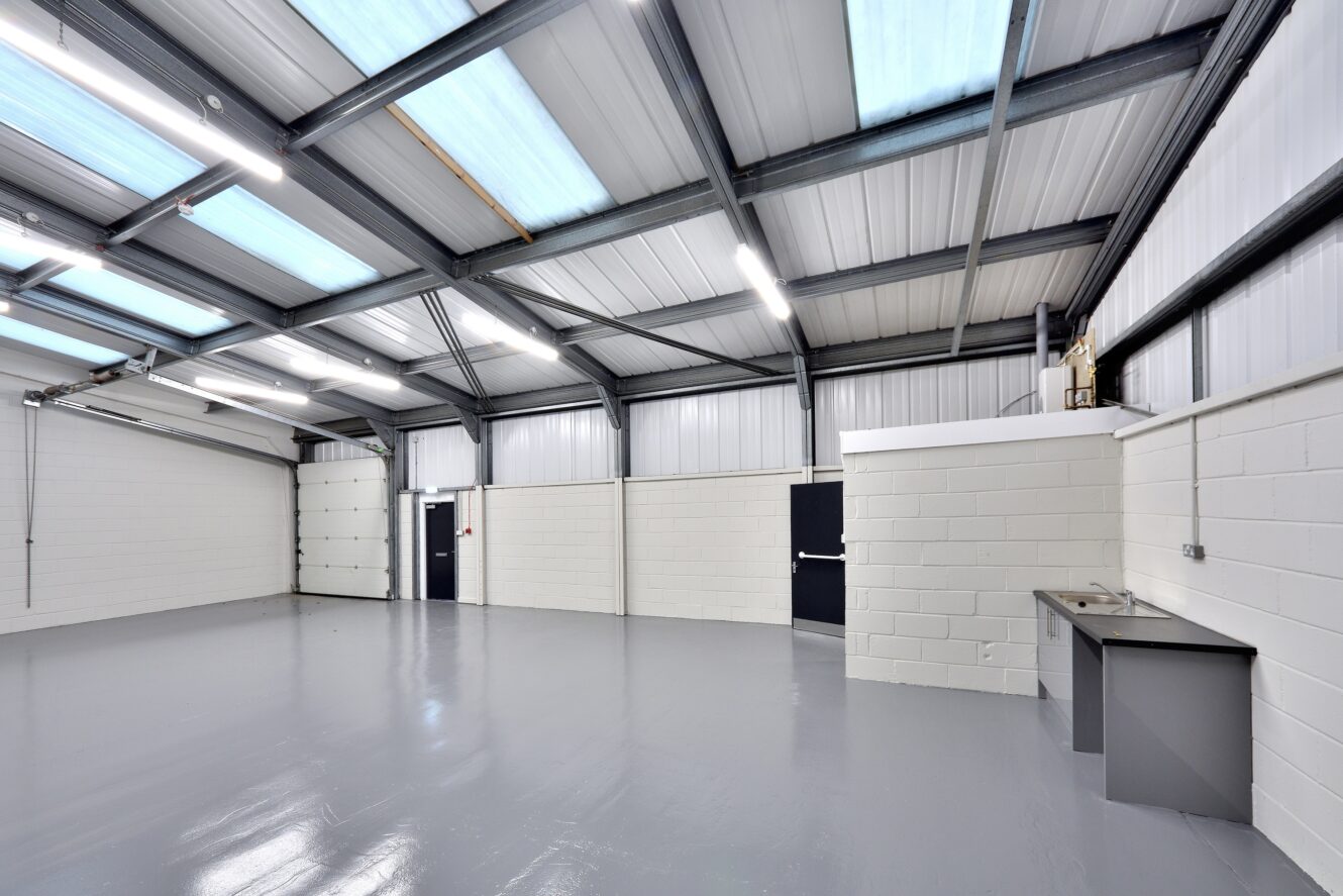 Empty industrial warehouse with white brick walls, grey painted floor, metal ceiling beams, skylights, and a small sink unit in the corner.