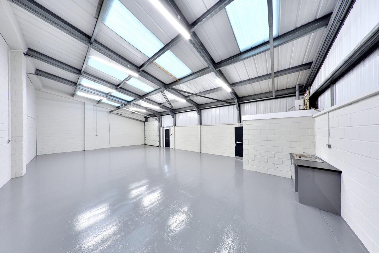 Interior of an empty industrial warehouse with white brick walls, a gray polished floor, skylights in the metal roof, and a small utility sink in the corner.