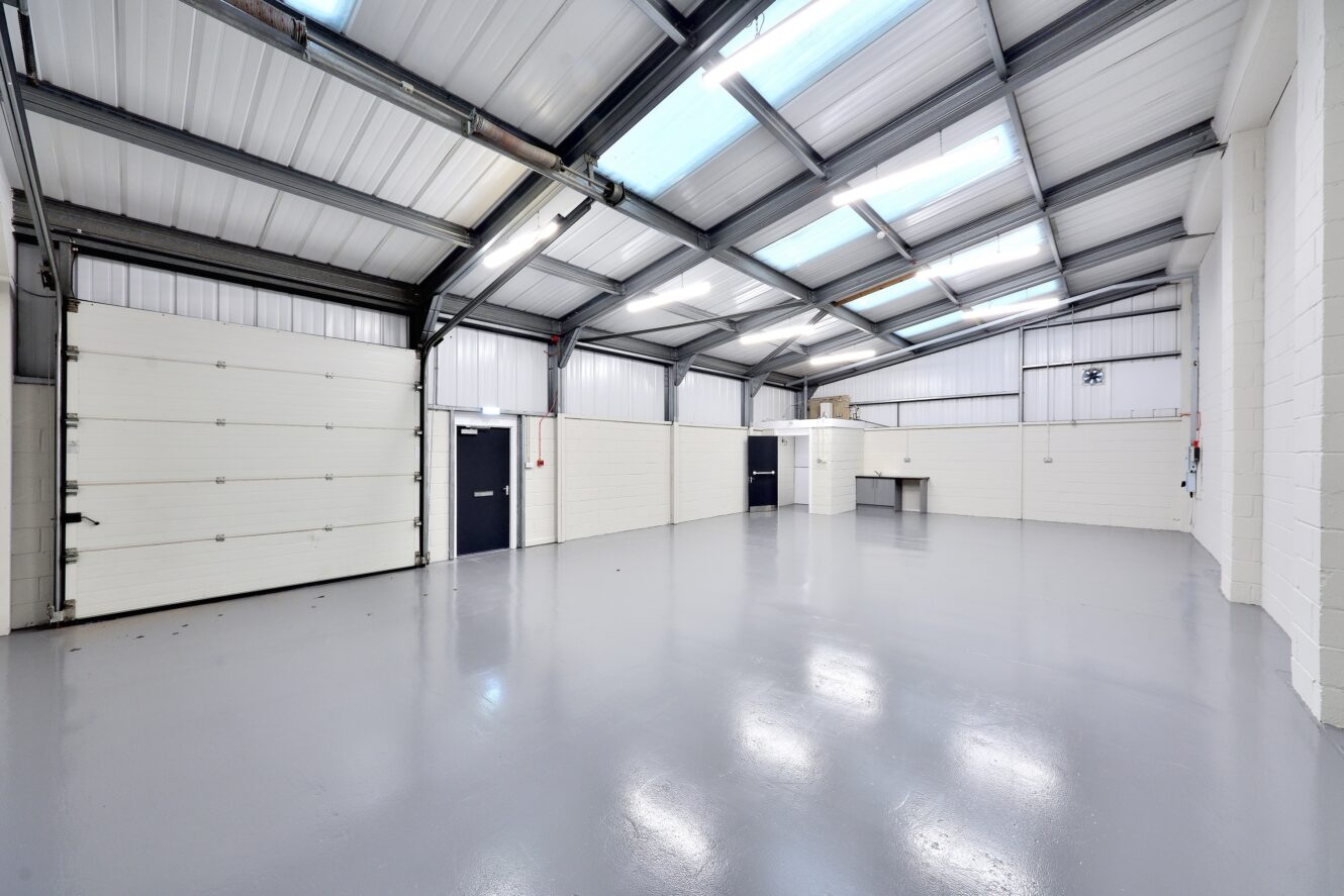 Interior of an empty industrial warehouse with white walls, polished gray floor, high ceiling, skylights, and large garage doors.