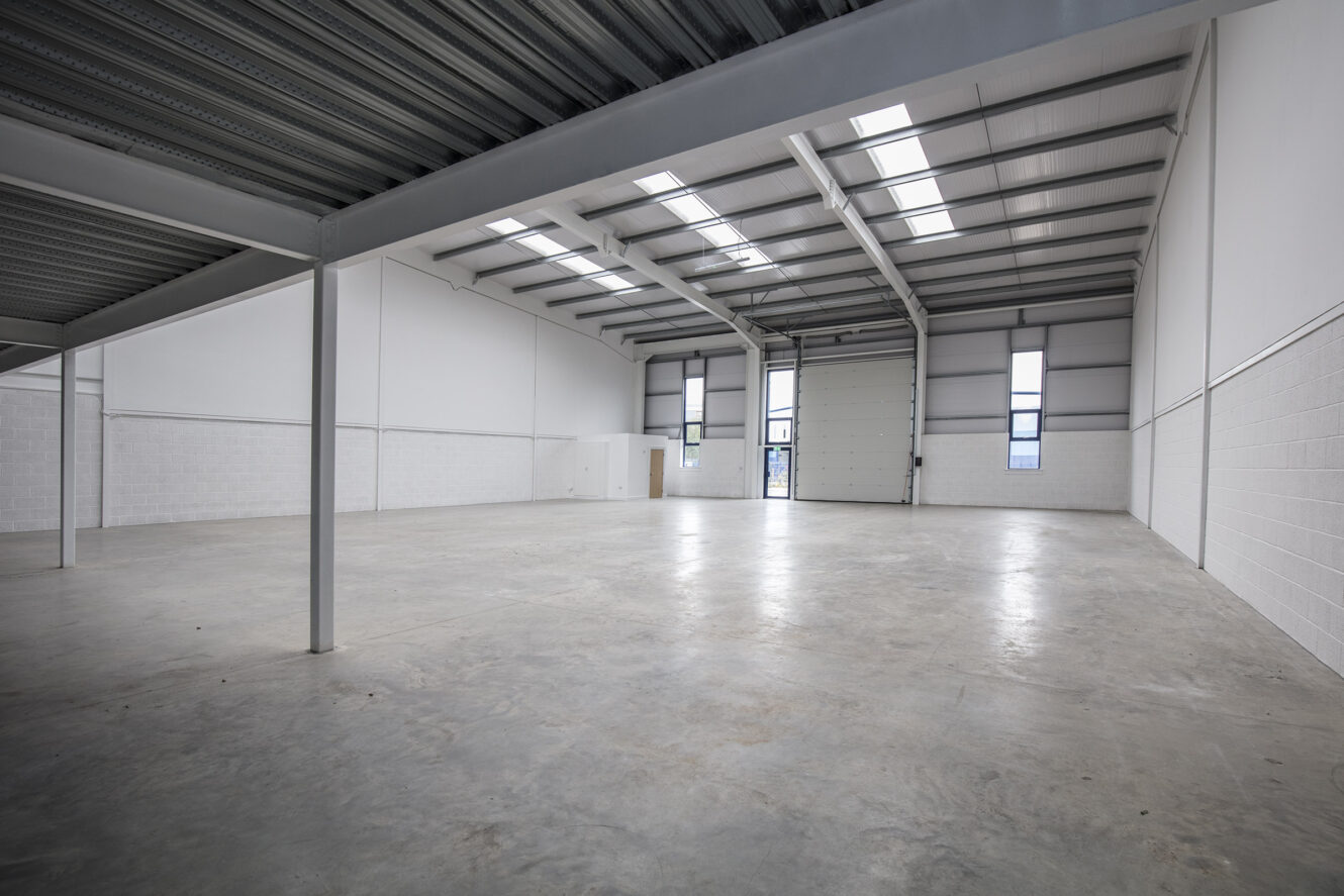 Interior of an empty industrial warehouse with concrete floor, high ceiling, large windows, and a roller shutter door.
