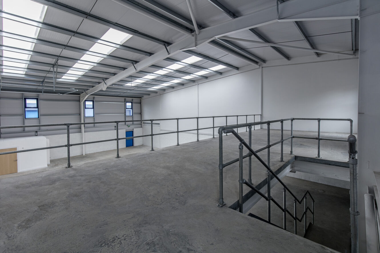Interior of an empty industrial warehouse mezzanine with concrete flooring, metal railings, and stairs; large windows provide natural light from above.