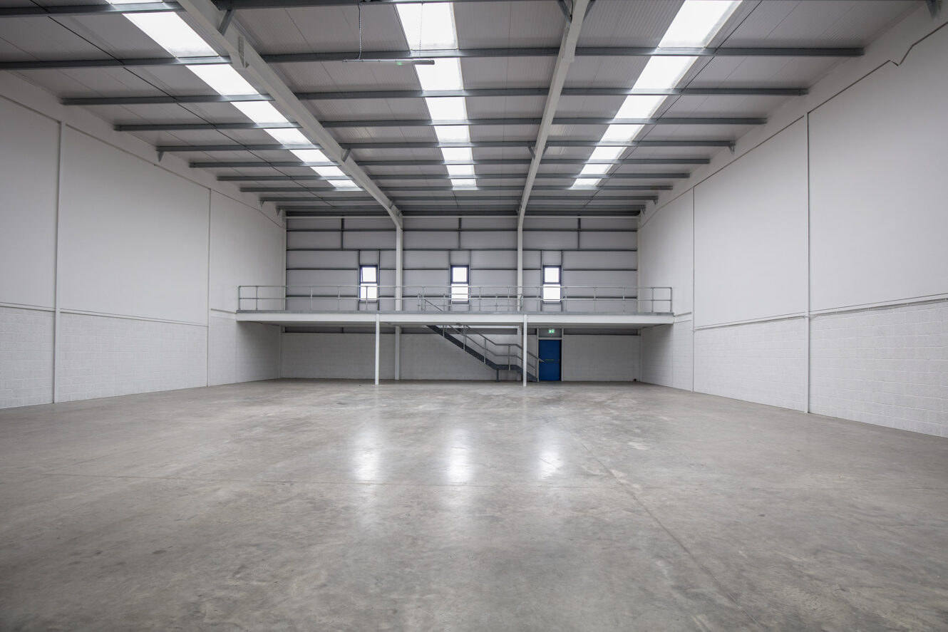 Empty industrial warehouse with concrete floor, high ceiling, mezzanine level, metal staircase, and rows of ceiling lights.