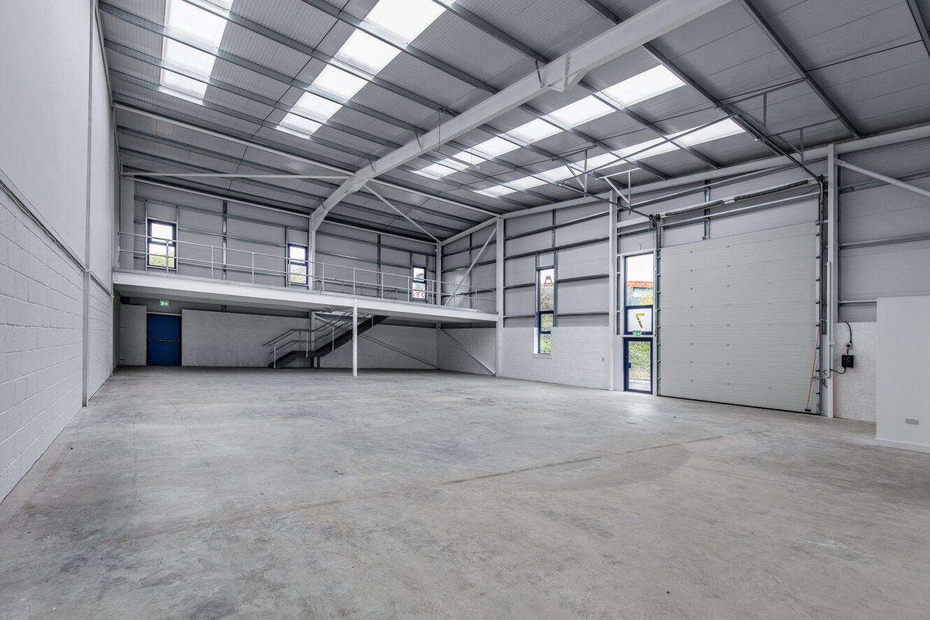 Spacious, empty industrial warehouse with concrete floor, high ceiling, metal beams, mezzanine level, staircase, and large roller door.