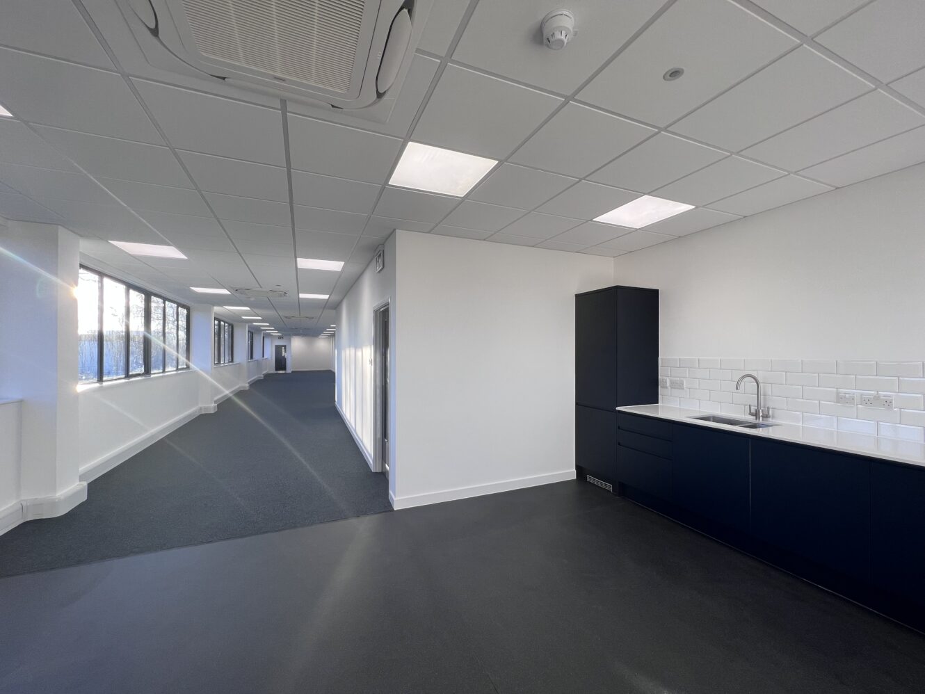 Modern office space with a kitchenette on the right, white walls, dark flooring, carpeted corridor, large windows, and overhead lighting.