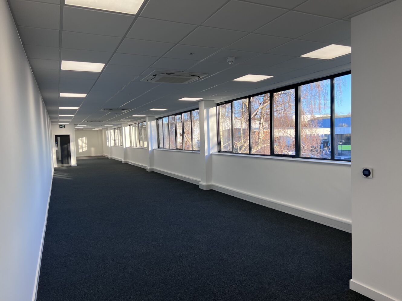 A long, empty office hallway with blue carpet, white walls, ceiling lights, and large windows letting in natural light.