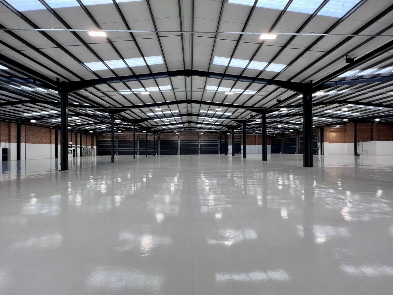 Interior of a large empty warehouse with polished floors, high ceilings, black steel beams, and brick walls, illuminated by overhead lights and skylights.