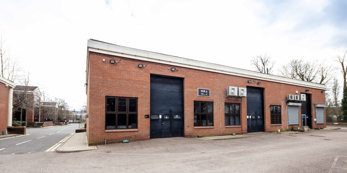 A row of brick industrial units with black doors, large windows, and air conditioning units mounted above, set in an empty parking area on a cloudy day.