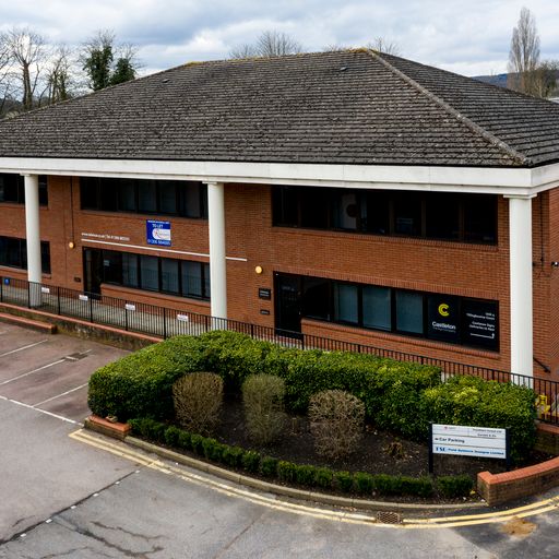 A two-story brick office building with large windows, surrounded by bushes and an empty parking lot, under a cloudy sky.