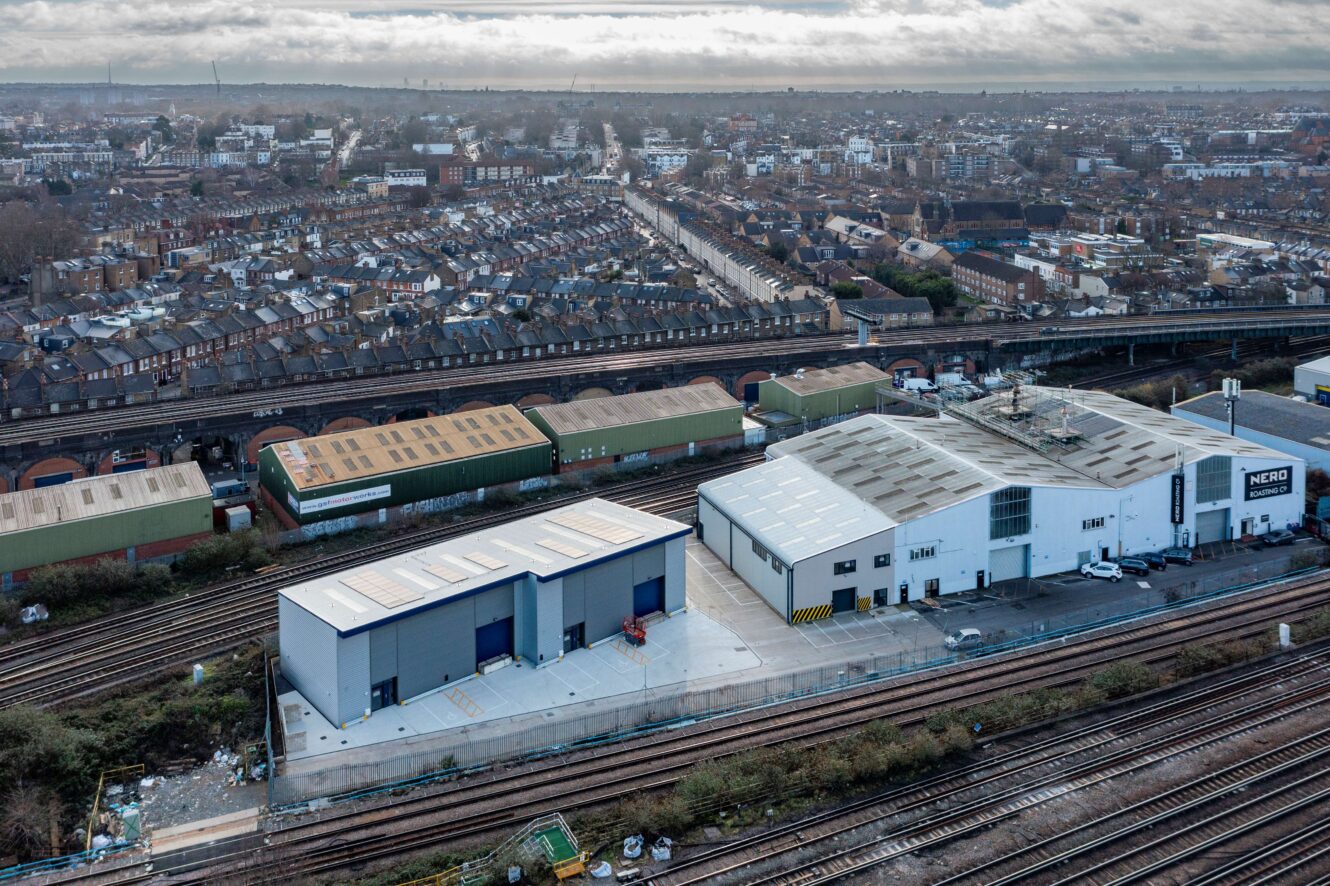 Aerial view of warehouses and industrial buildings beside railway tracks, with residential houses and a cityscape in the background under a cloudy sky.