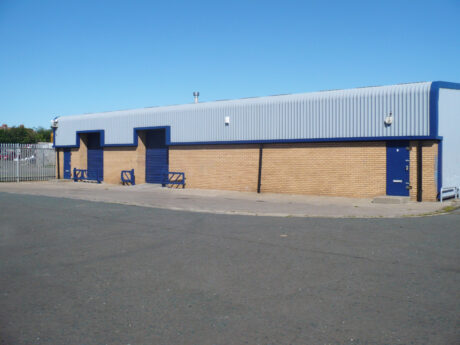 A single-story industrial building with brick walls and blue roller doors, set on a large paved area under a clear blue sky.