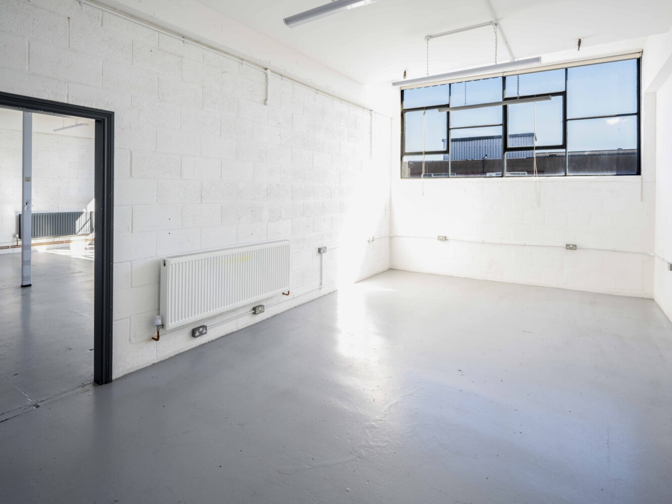 An empty, white-walled industrial room with a large window letting in sunlight, grey floor, and a radiator on one wall.