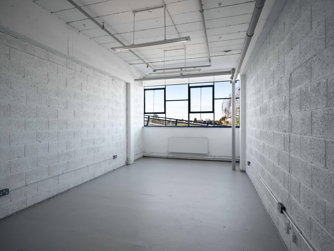 Empty white-walled room with concrete floor, exposed pipes, and large windows letting in natural light.
