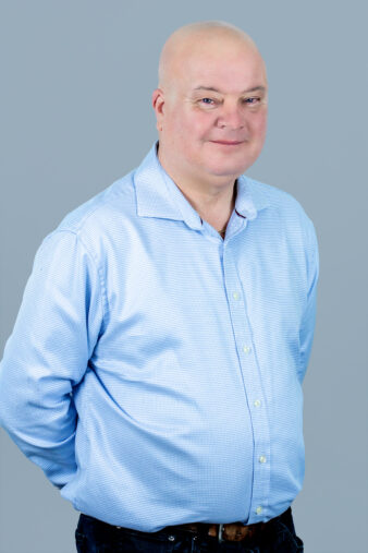 A bald man wearing a light blue button-up shirt stands with his hands behind his back against a plain gray background.