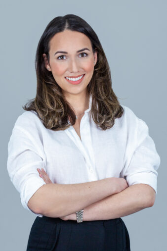 A woman with shoulder-length brown hair, wearing a white shirt and black pants, stands with arms crossed and smiles against a plain gray background.
