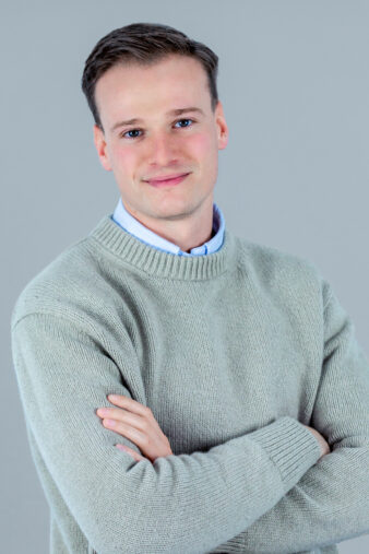 A young man with short brown hair, wearing a light gray sweater over a collared shirt, stands with arms crossed and a neutral expression against a plain gray background.