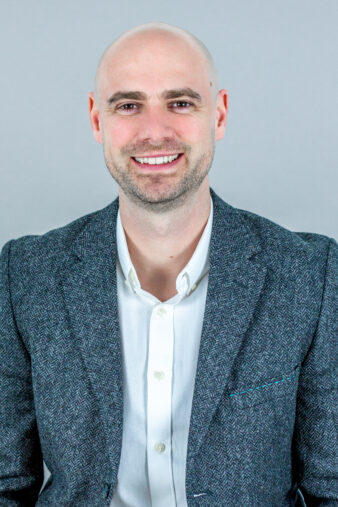 A bald man in a grey blazer and white shirt smiles at the camera against a plain light grey background.
