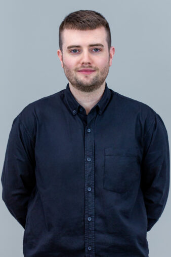 A man with short brown hair and a trimmed beard, wearing a black button-up shirt, standing against a plain gray background.