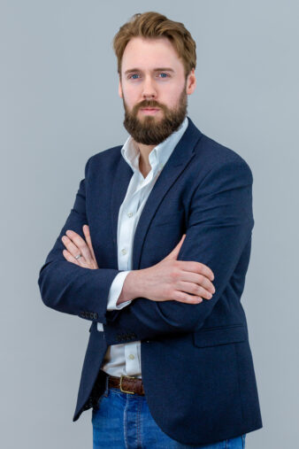 A man with a beard and mustache stands with arms crossed, wearing a navy blazer, white shirt, and blue jeans against a plain light gray background.