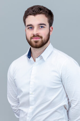A young man with a beard and short brown hair stands against a plain light gray background, wearing a white button-down shirt.