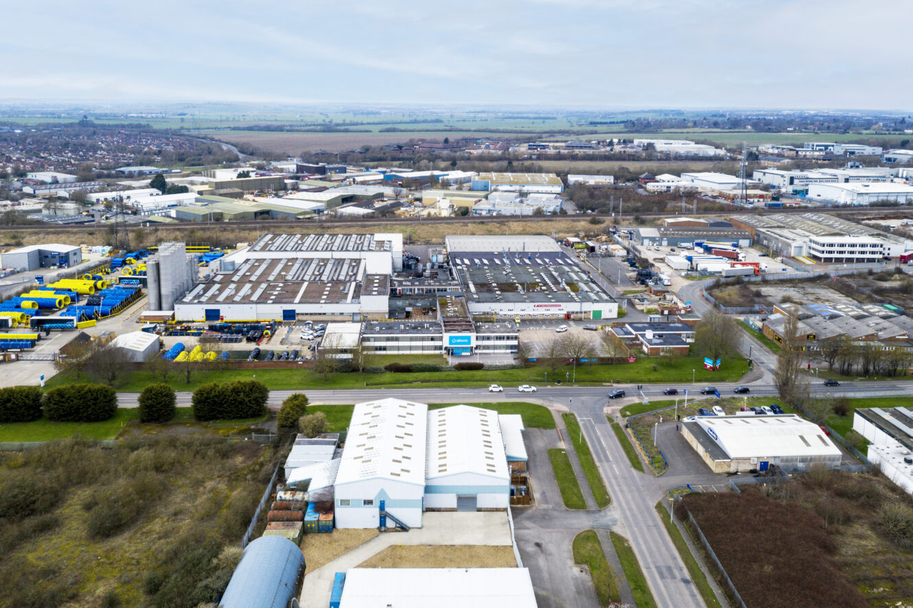 Aerial view of an industrial estate with large warehouses, factories, parking lots, and roads surrounded by some greenery and distant residential areas.