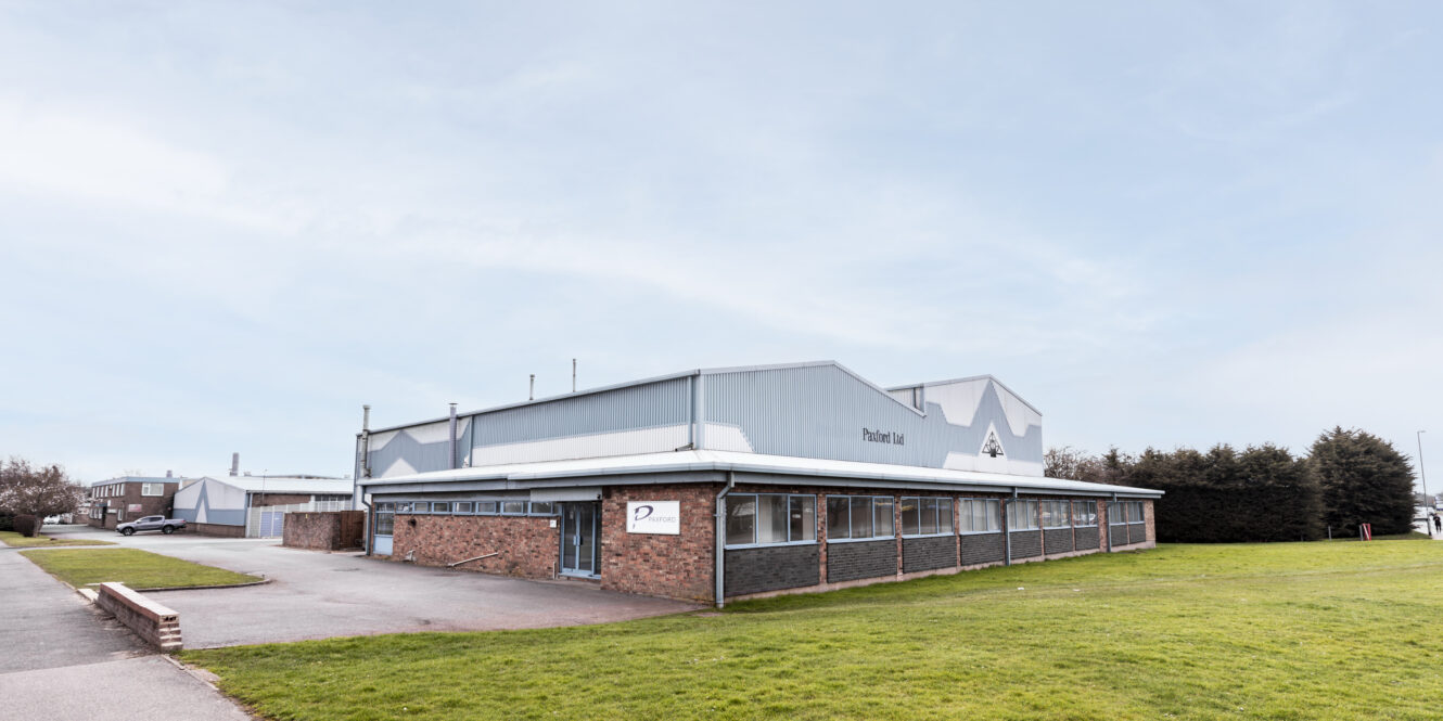 A single-story industrial building with a brick and metal exterior sits next to a paved driveway and a grassy area under a clear sky.