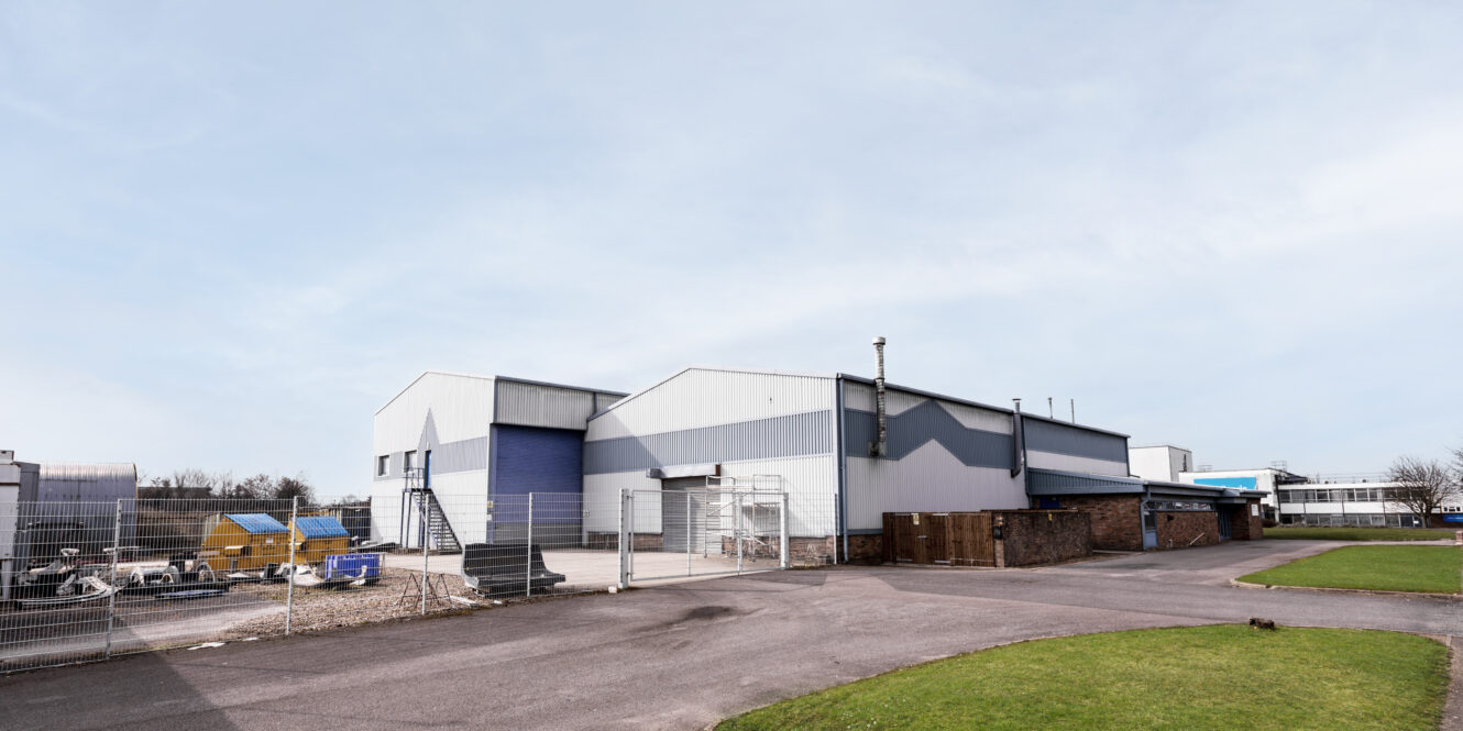 Industrial warehouse with metal siding and blue accents, surrounded by fencing and outdoor storage containers, under a clear sky.