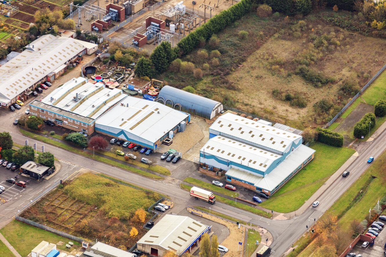 Aerial view of an industrial estate with several warehouse buildings, parking lots, a few vehicles, and adjacent green spaces.