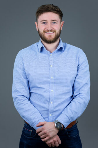 A man with short brown hair and a beard, wearing a light blue button-up shirt and jeans, stands against a plain grey background, smiling with his hands clasped in front.