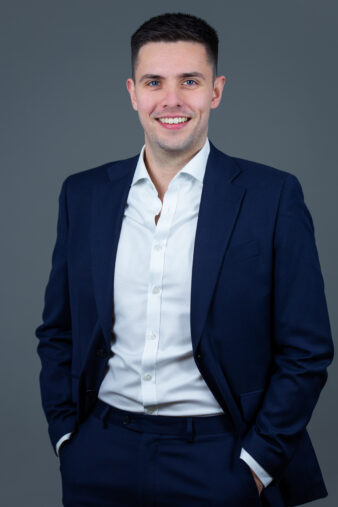 A man in a navy suit and white dress shirt stands against a plain gray background, smiling with his hands in his pockets.
