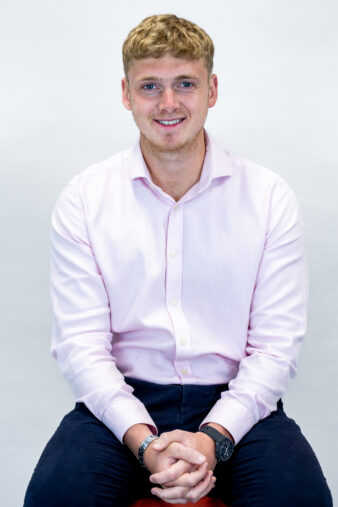 A young man with short blond hair wearing a light pink shirt and dark trousers sits on a stool against a plain white background, smiling at the camera.