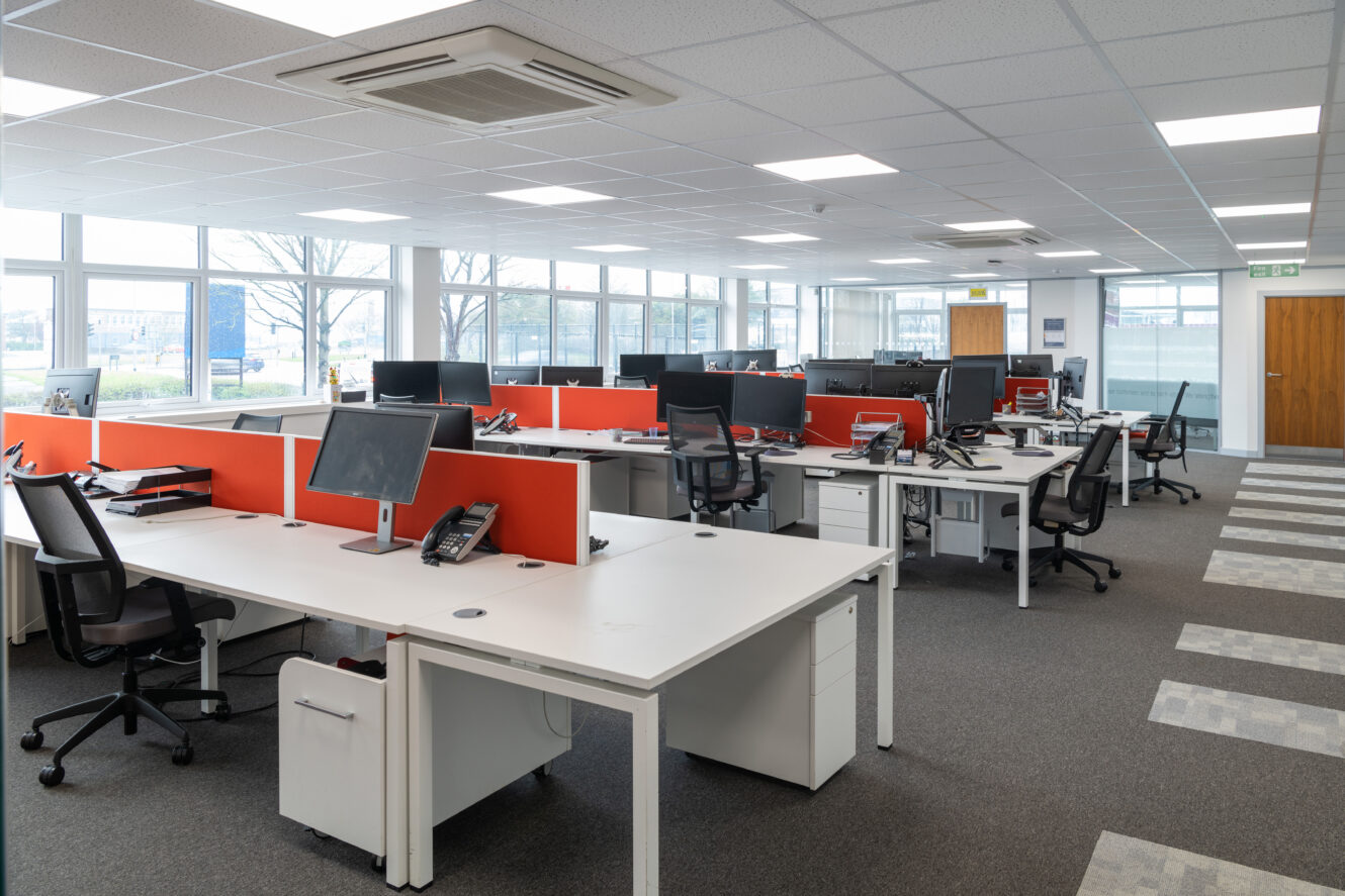 Modern open-plan office with multiple desks, computers, and chairs. The space is well-lit with large windows and has red desk dividers. No people are present.