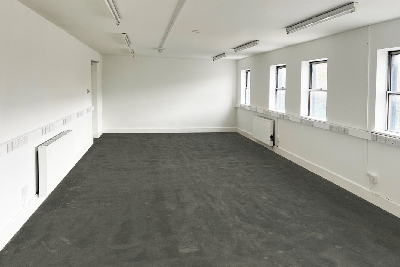 Empty room with plain white walls, multiple ceiling lights, three windows on one side, wall-mounted heaters, and a dark carpeted floor.