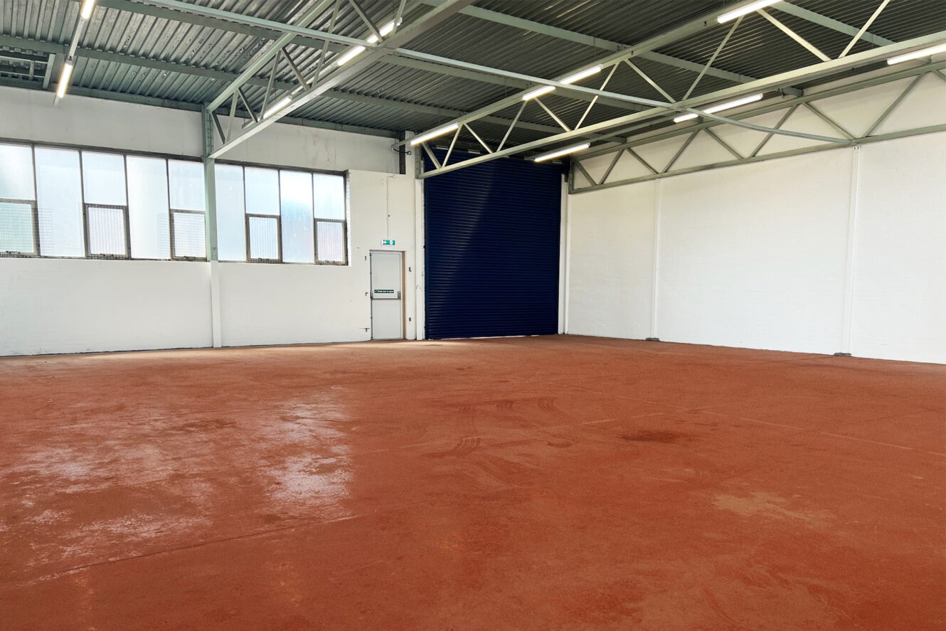 An empty industrial warehouse with red flooring, white walls, large windows, exposed beams, and a blue roller shutter door.