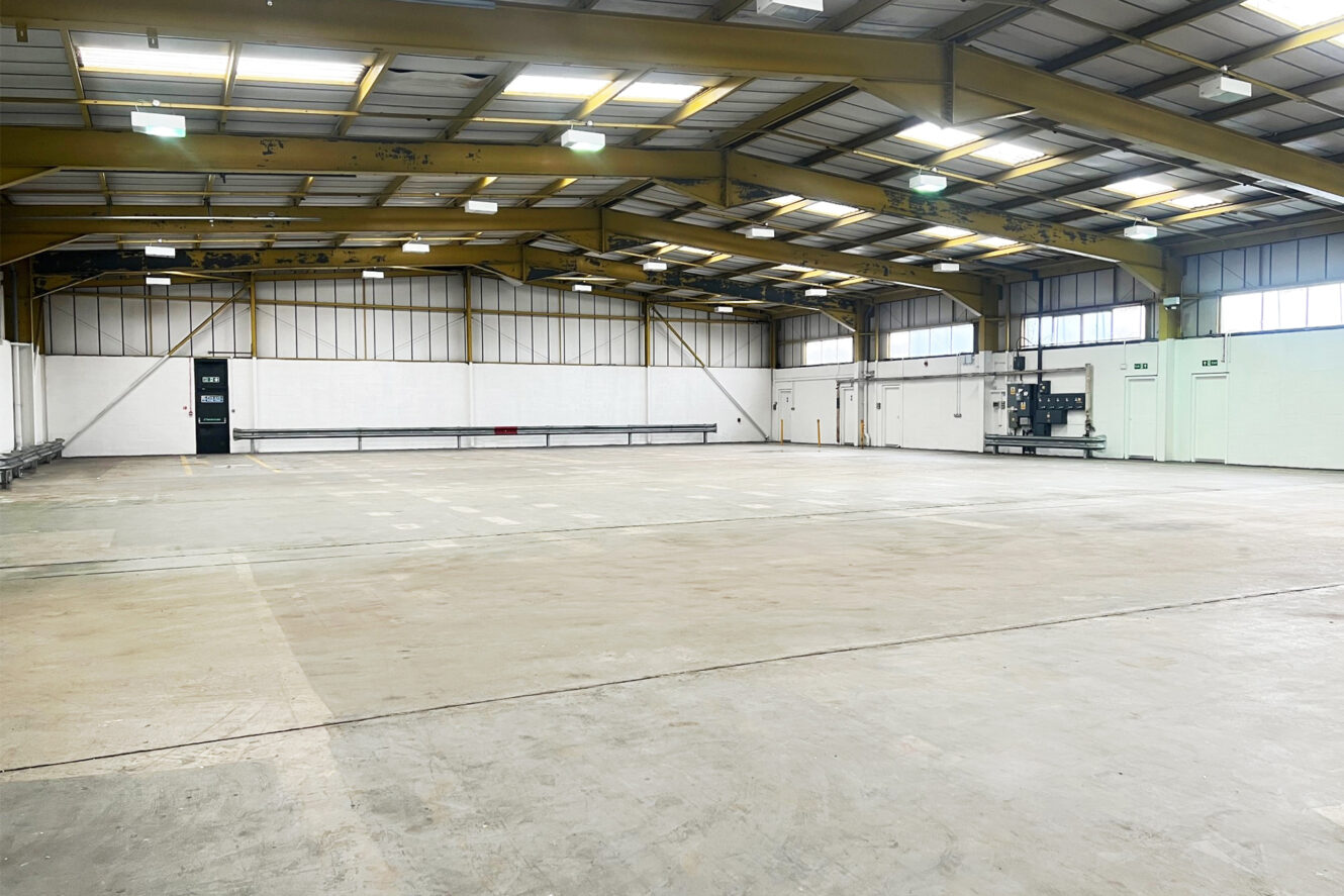 Large empty industrial warehouse with high ceilings, exposed steel beams, and overhead lighting. A forklift is parked near the white walls on the right side.