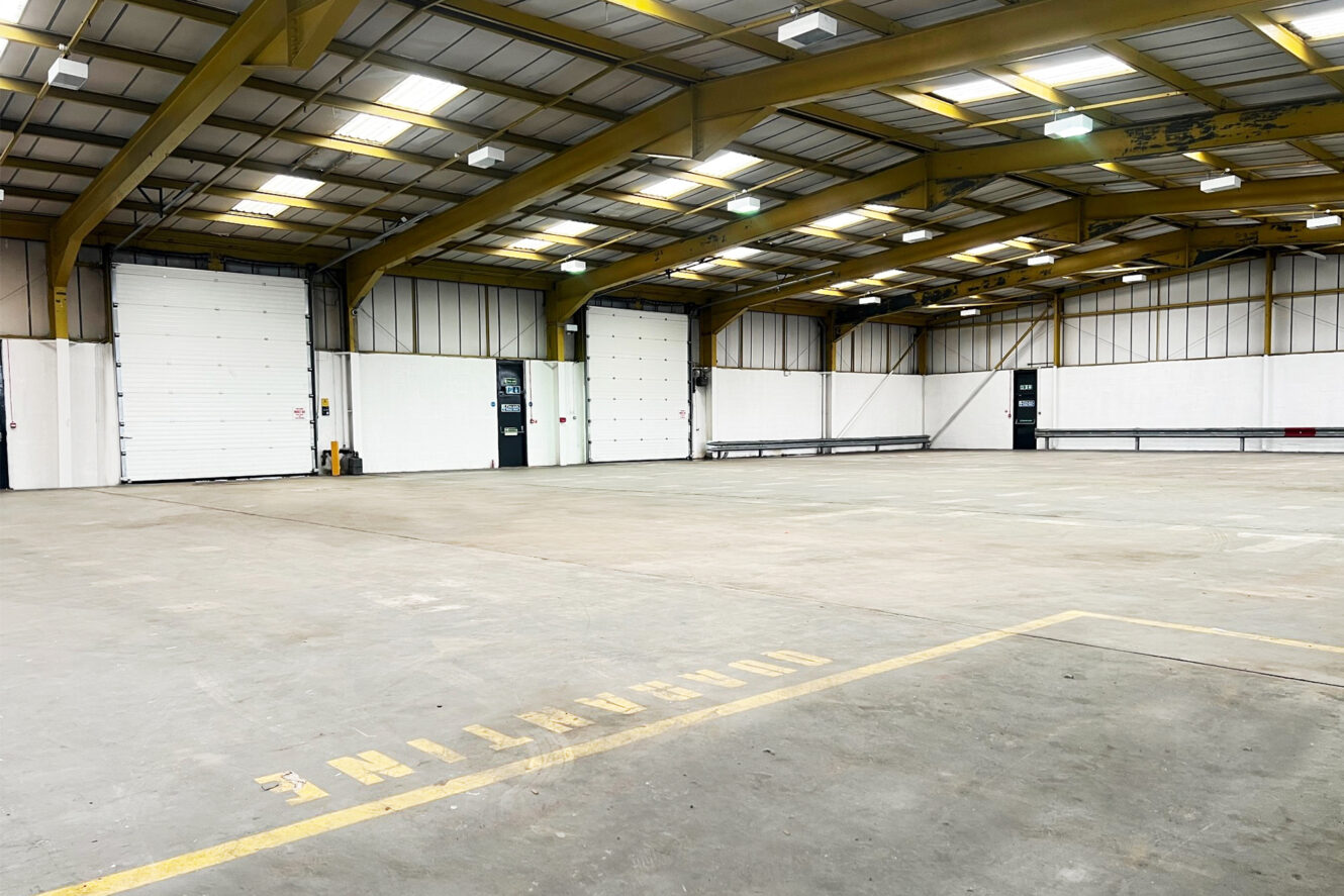 Large empty warehouse interior with high ceiling, yellow steel beams, concrete floor, and several loading bay doors along the walls.