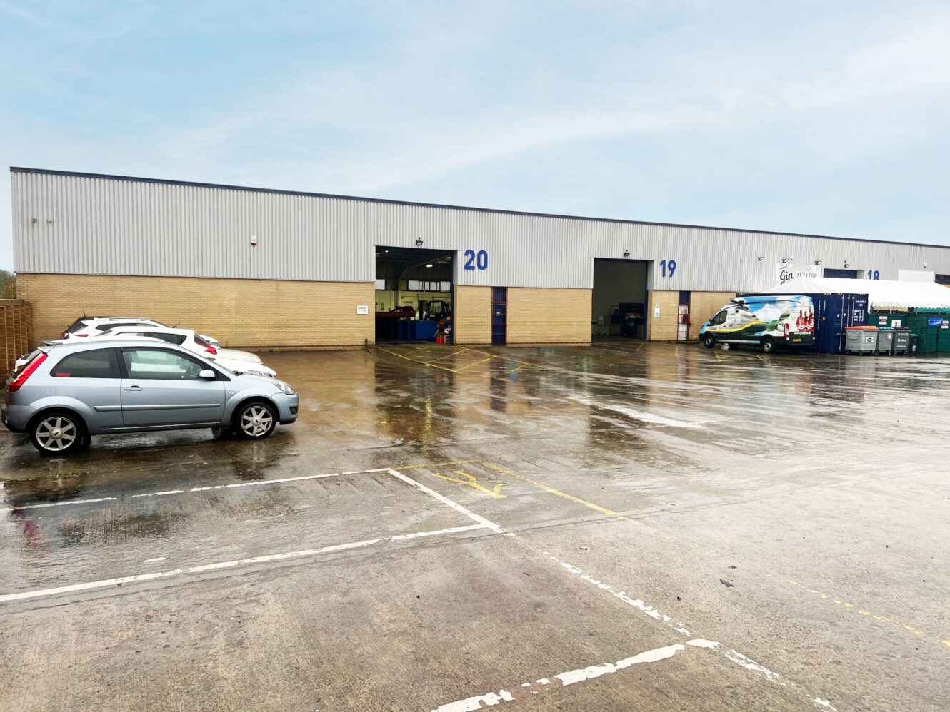 A wet parking lot outside a large warehouse with unit numbers 19 and 20. Several parked cars and a van are visible near open loading bays.