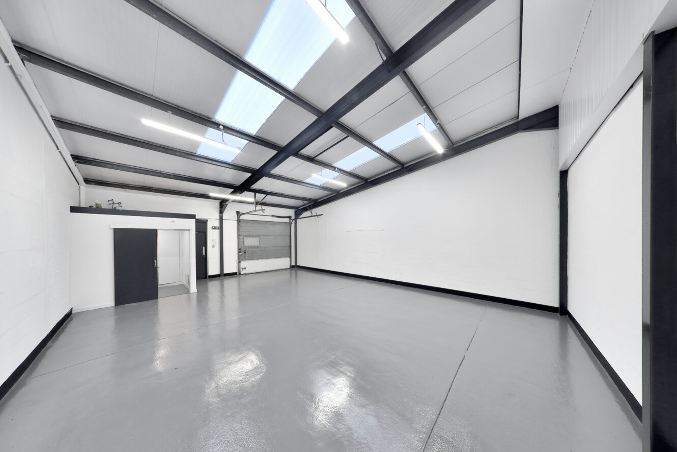 Empty industrial warehouse with white walls, gray floor, high ceiling, skylights, fluorescent lights, a gray door, and a large roller shutter door.