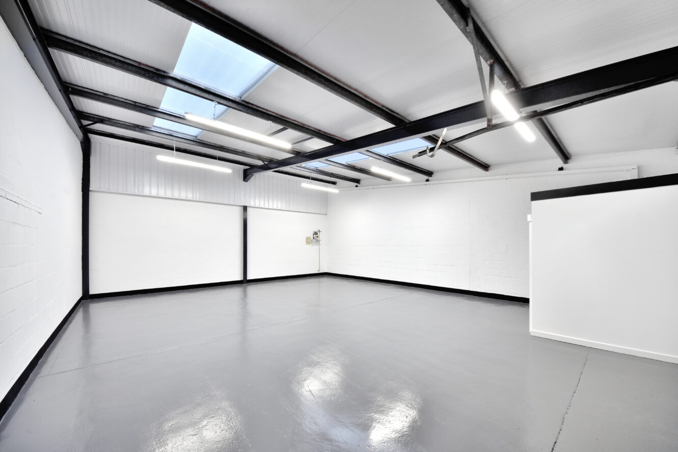 Empty industrial-style room with white walls, exposed black ceiling beams, polished gray floor, and skylights letting in natural light.