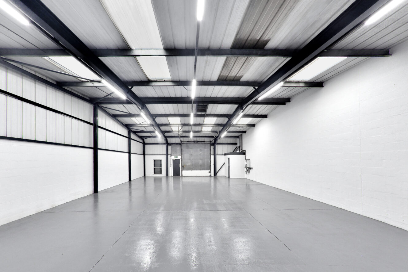 Empty industrial warehouse interior with white painted walls, high ceiling, exposed beams, gray concrete floor, and fluorescent lighting. A metal door and staircase are visible at the back.