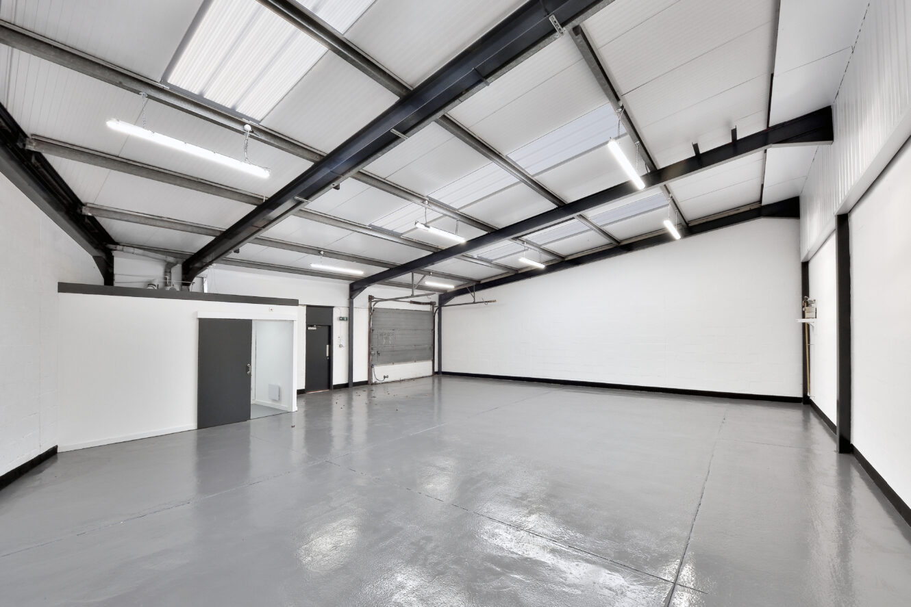 Empty industrial warehouse with white walls, high ceiling, exposed beams, overhead lights, a small office, and a large roller shutter door.