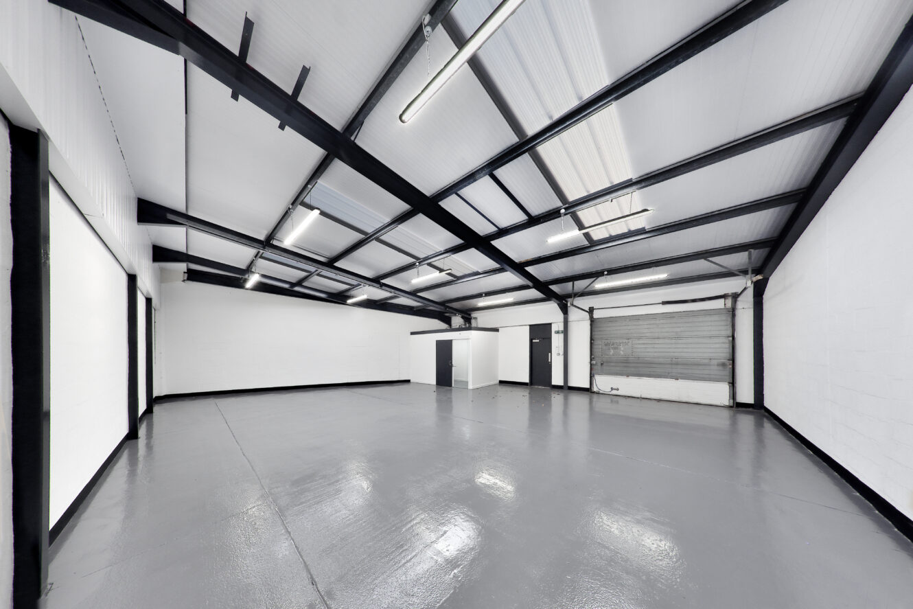 Empty industrial warehouse with white walls, a polished concrete floor, exposed ceiling beams, fluorescent lighting, and a large roller shutter door.