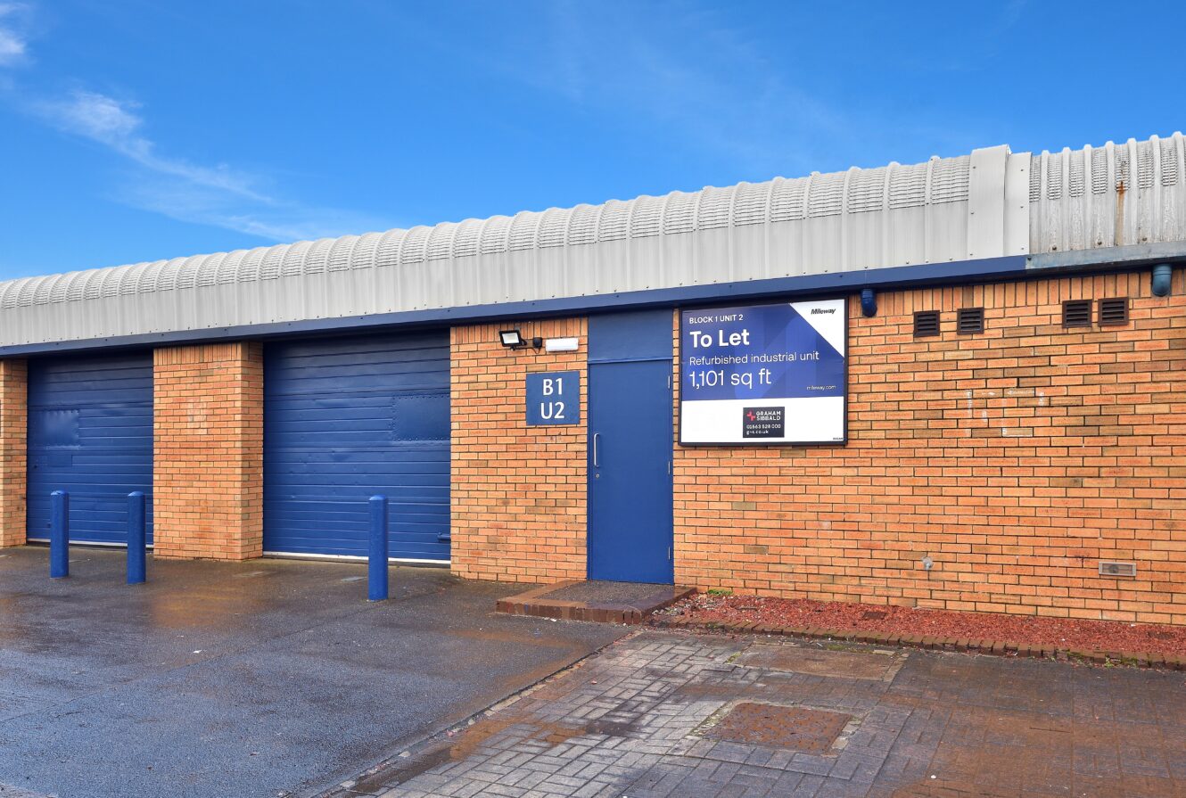 A brick industrial unit with blue doors and a sign advertising 1,101 sq ft available to let, under a clear blue sky.