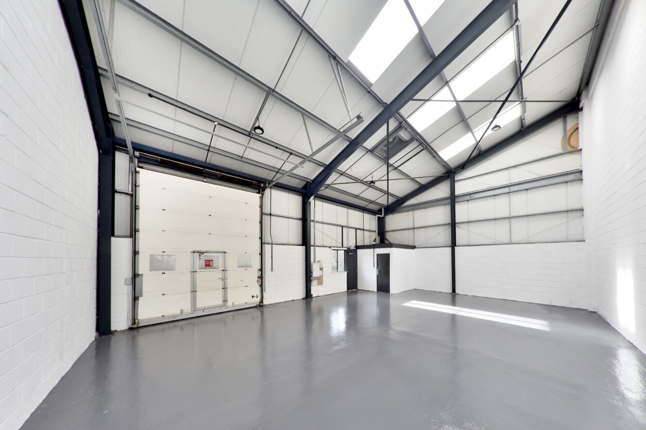 Empty industrial warehouse with white painted brick walls, high ceiling, large overhead door, and gray polished concrete floor. Natural light enters through skylights and windows.