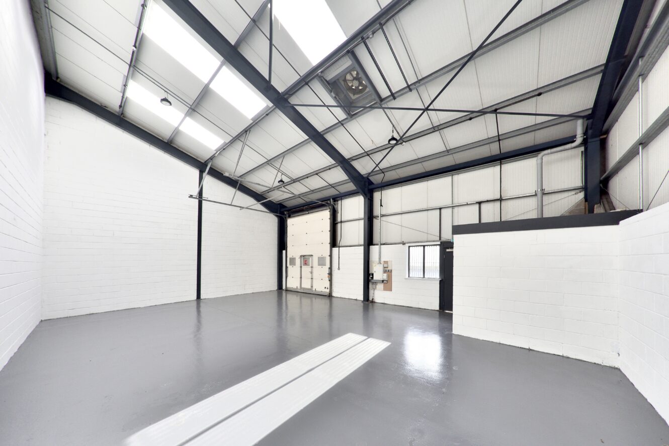 Empty industrial warehouse with high ceilings, white walls, large overhead door, gray polished floor, and exposed metal beams. Natural light enters through roof panels.