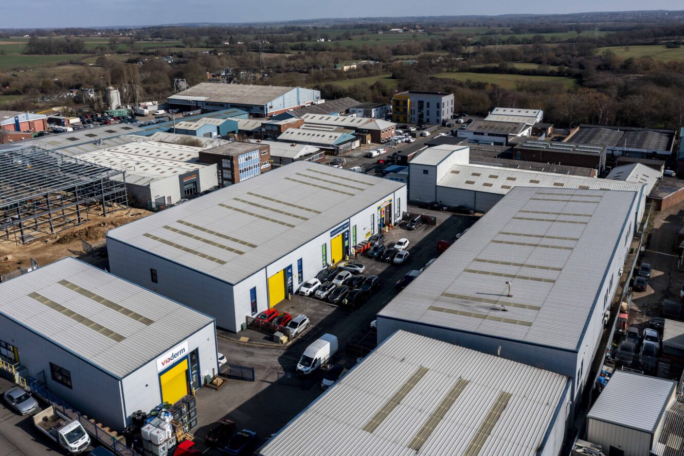 Aerial view of an industrial estate with multiple large warehouse buildings, parked cars, and surrounding countryside visible in the background.