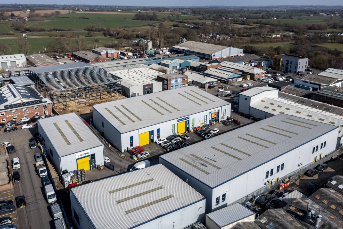 Aerial view of an industrial estate with multiple warehouses, parked cars, and surrounding countryside in the background.